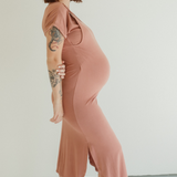 A pregnant woman stands barefoot on a white backdrop wearing the Chekoh Baby Journey Dress in the colour Dusk - a neutral dusky pink tone.