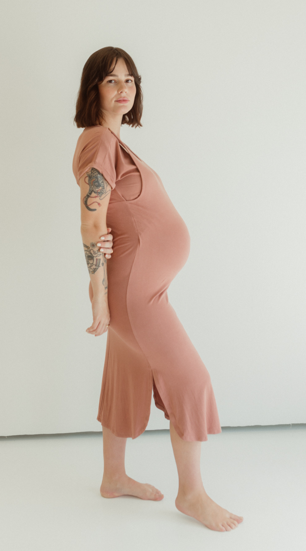 A pregnant woman stands barefoot on a white backdrop wearing the Chekoh Baby Journey Dress in the colour Dusk - a neutral dusky pink tone.