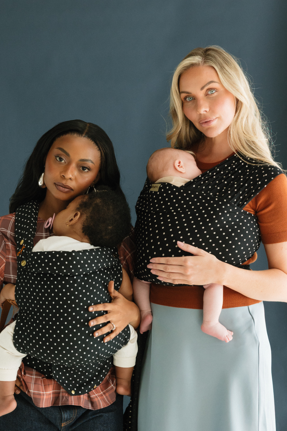 Two mothers stand side by side, one wearing the new Clip Carrier and one wearing the new Stretchy Wrap Carrier in the Polka Dot print by Chekoh Baby. Both baby's look sleepy and rested.