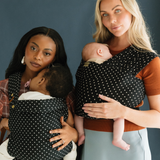 Two mothers stand side by side, one wearing the new Clip Carrier and one wearing the new Stretchy Wrap Carrier in the Polka Dot print by Chekoh Baby. Both baby's look sleepy and rested.