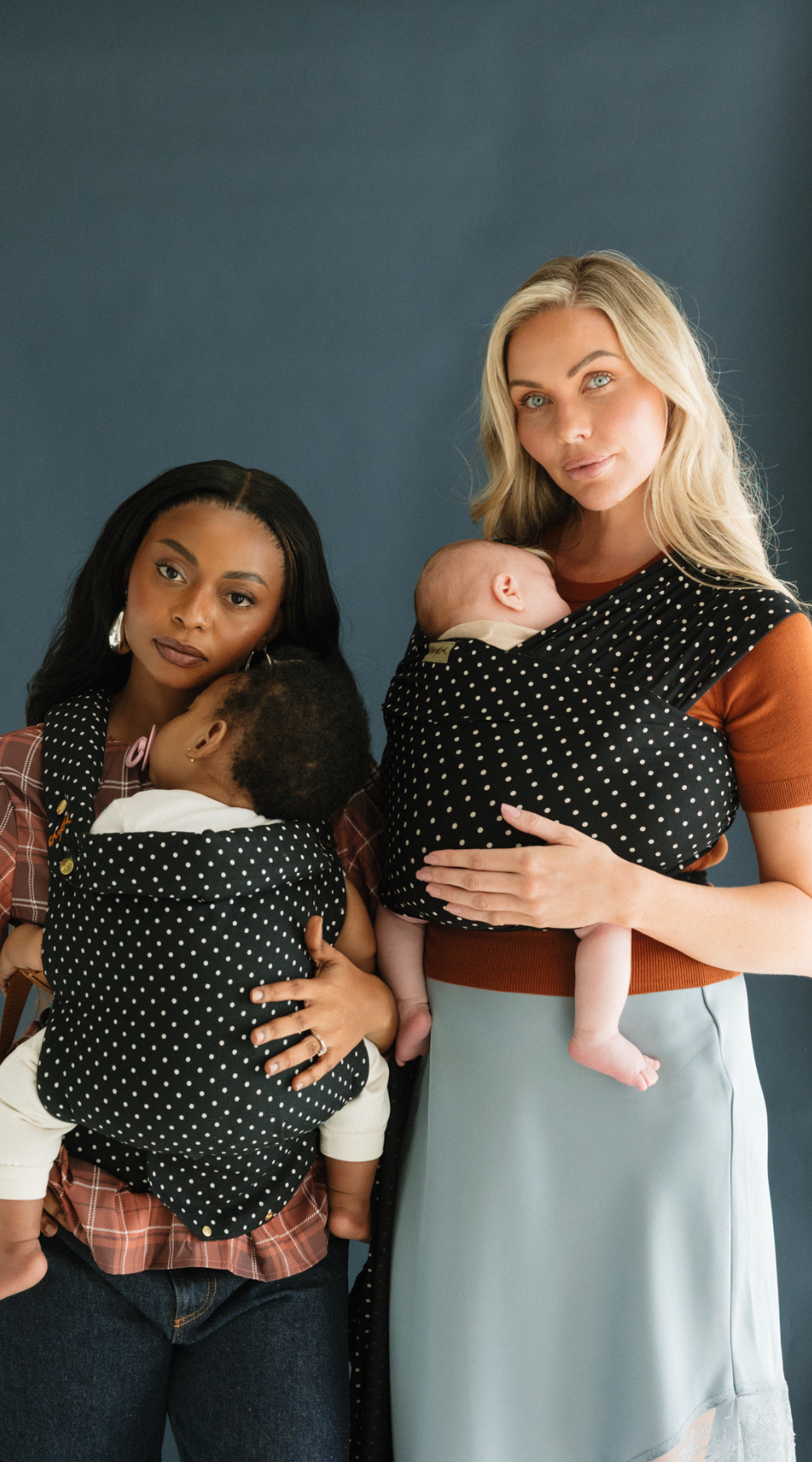 Two mothers stand side by side, one wearing the new Clip Carrier and one wearing the new Stretchy Wrap Carrier in the Polka Dot print by Chekoh Baby. Both baby's look sleepy and rested.