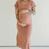 A pregnant woman holds her belly while standing barefoot and comfortable against a white backdrop. She is wearing the Dusk Journey Dress by Chekoh Baby.