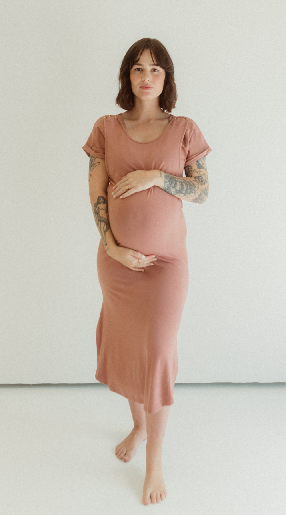 A pregnant woman holds her belly while standing barefoot and comfortable against a white backdrop. She is wearing the Dusk Journey Dress by Chekoh Baby.