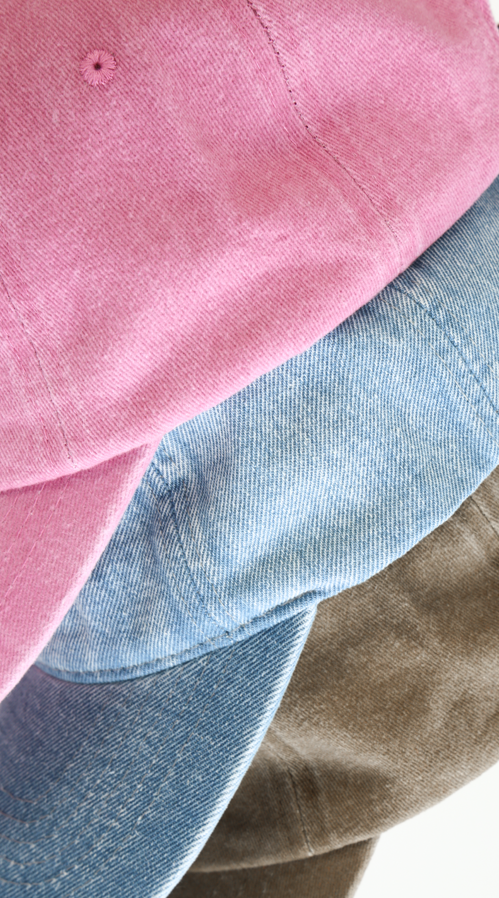 Stack of three baseball caps in Pink, Lightwash Denim, and Bone on a white background.