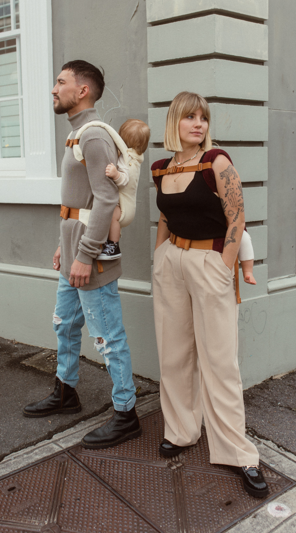 Man and woman, wearing their babies in a back carry using the Chekoh Chest Strap for added support.