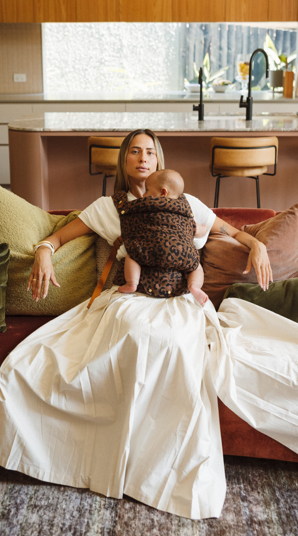 Woman in white dress is wearing the Leopard Clip Carrier 2.0 by Chekoh Baby, sat on a couch soaking up the closeness between the two of them.