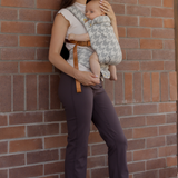 Woman holding a baby against a brick wall. Mom is wearing activewear, a Chekoh Cap and the Hudson houndstooth Clip Carrier - a gray houndstooth woven print.