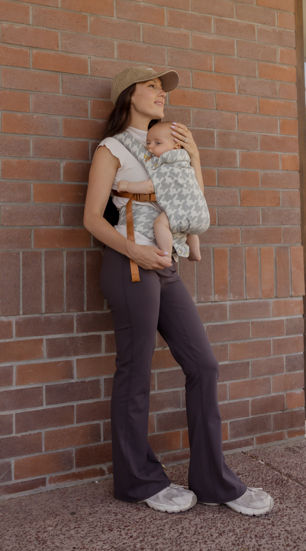 Woman holding a baby against a brick wall. Mom is wearing activewear, a Chekoh Cap and the Hudson houndstooth Clip Carrier - a gray houndstooth woven print.