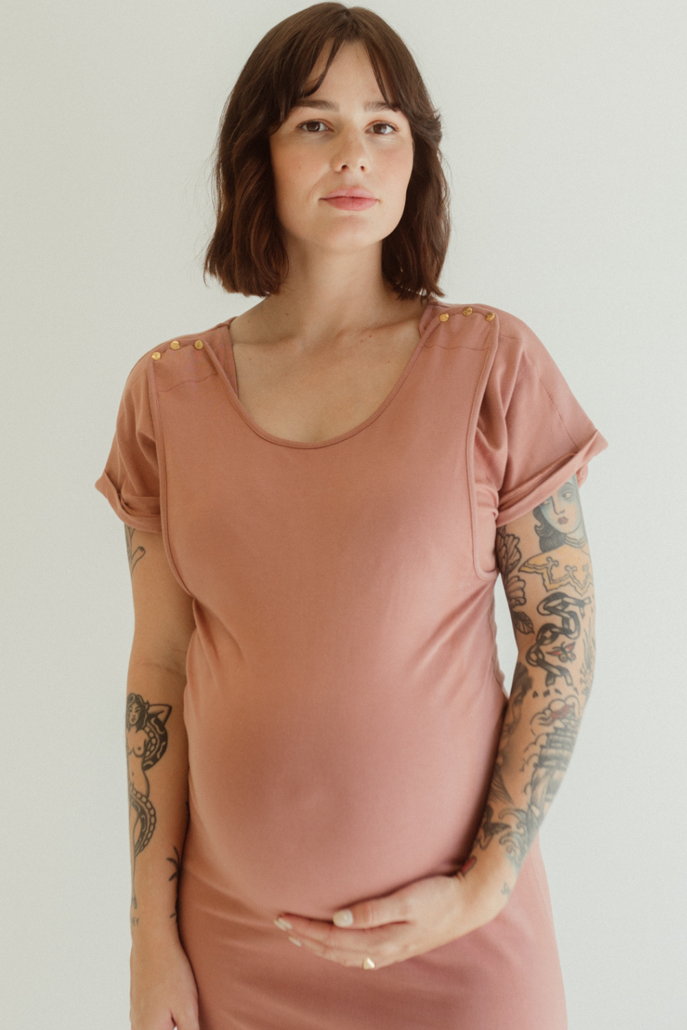 A pregnant mother wears the Dusk Journey Dress by Chekoh Baby. She is holding her pregnant belly and the dress is a soft dusky pink tone.