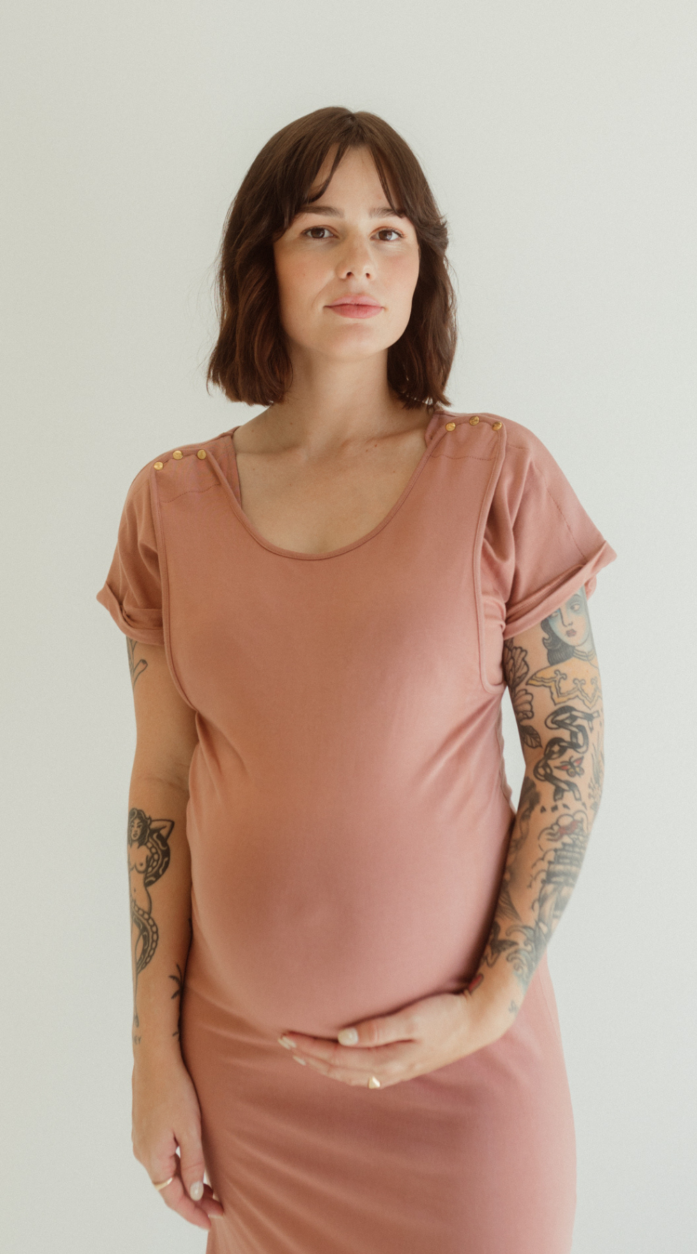 A pregnant mother wears the Dusk Journey Dress by Chekoh Baby. She is holding her pregnant belly and the dress is a soft dusky pink tone.