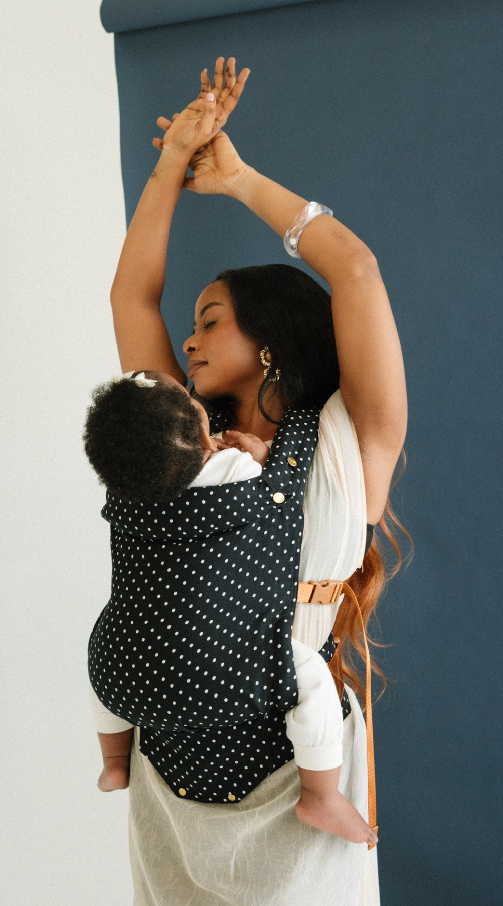 A woman holds her baby close and demonstrates that hands-free feeling you get when babywearing. She has her arms in the air and baby is looking up at her from the comfort of a Chekoh Baby Clip Carrier in Polka Dot.