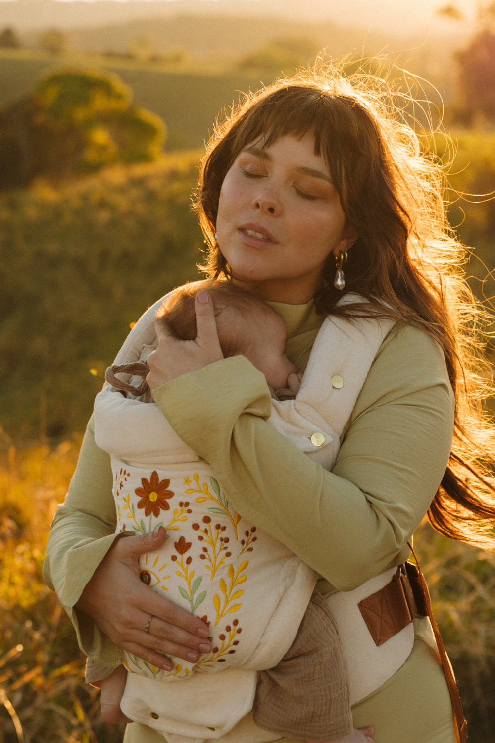 A mother and baby share a moment in a field wearing the vintage white cotton velvet Juni Clip. Featuring a stunning floral seeing eye embroidered design.