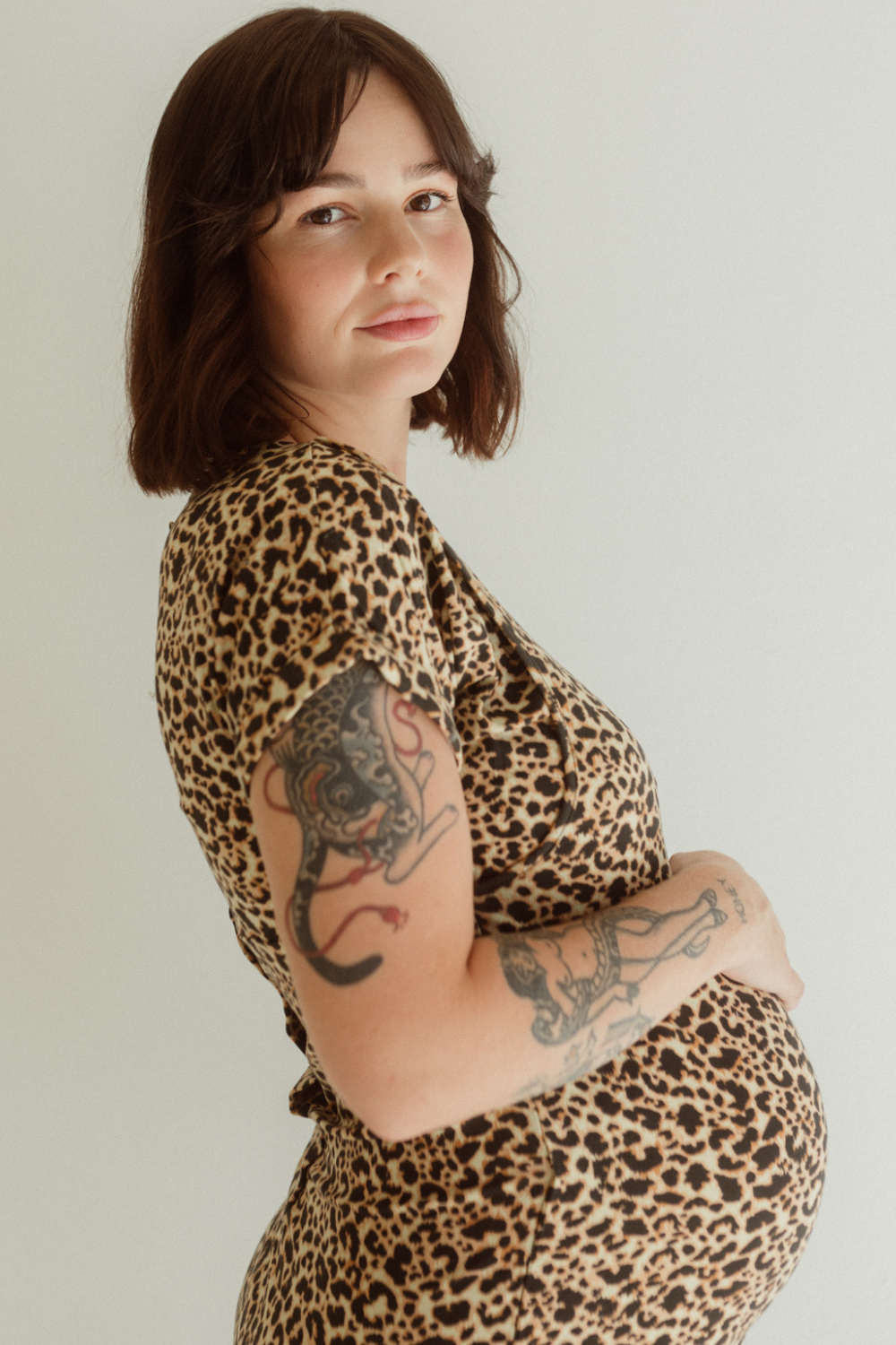 A pregnant woman wears the Leopard Print Journey Dress by Chekoh and holds her belly with one hand.