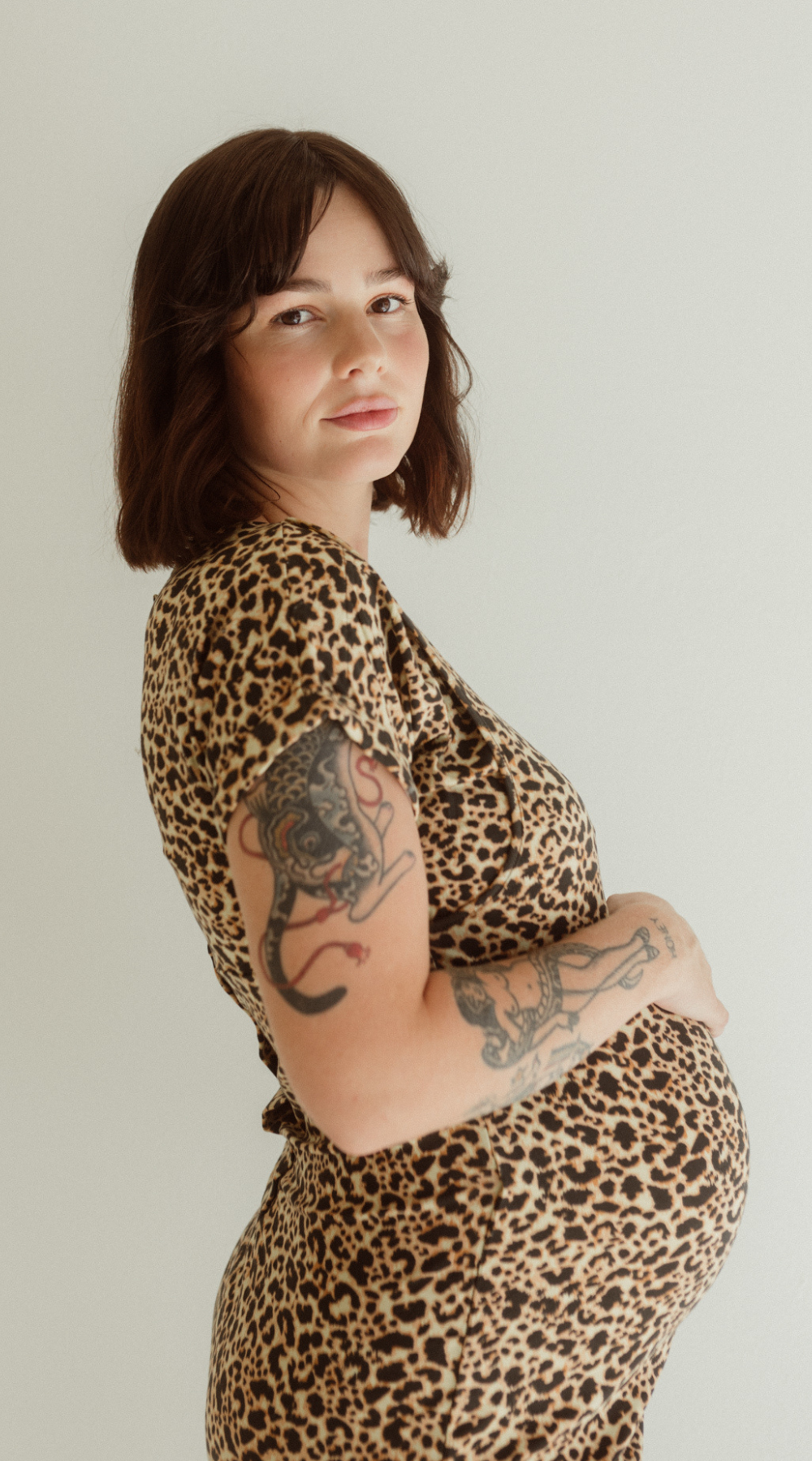 A pregnant woman wears the Leopard Print Journey Dress by Chekoh and holds her belly with one hand.