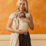 An Australian Mum is standing on a bright orange and yellow backdrop, smiling, while holding her newborn close in a Chekoh Baby Ring Sling in the shade Dune (a soft sandy neutral beige tone).