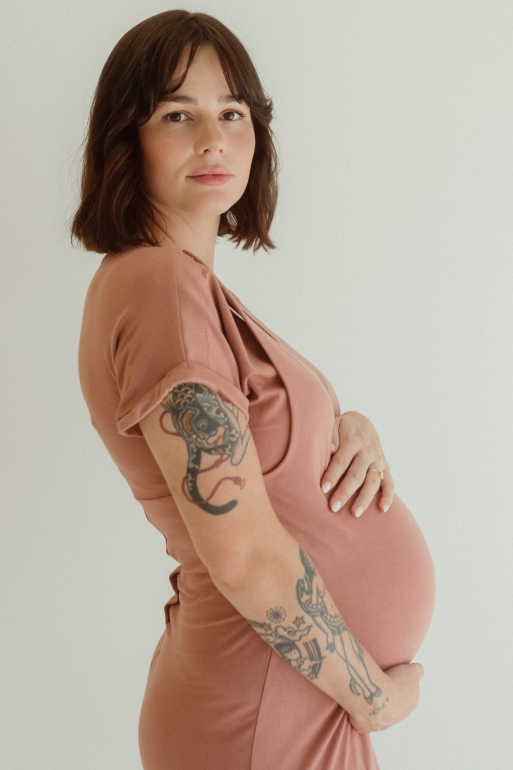 A pregnant mom holds her belly while wearing the Chekoh Journey Dress in the color Dusk - a dusky rose pink tone.