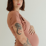 A pregnant mom holds her belly while wearing the Chekoh Journey Dress in the color Dusk - a dusky rose pink tone.