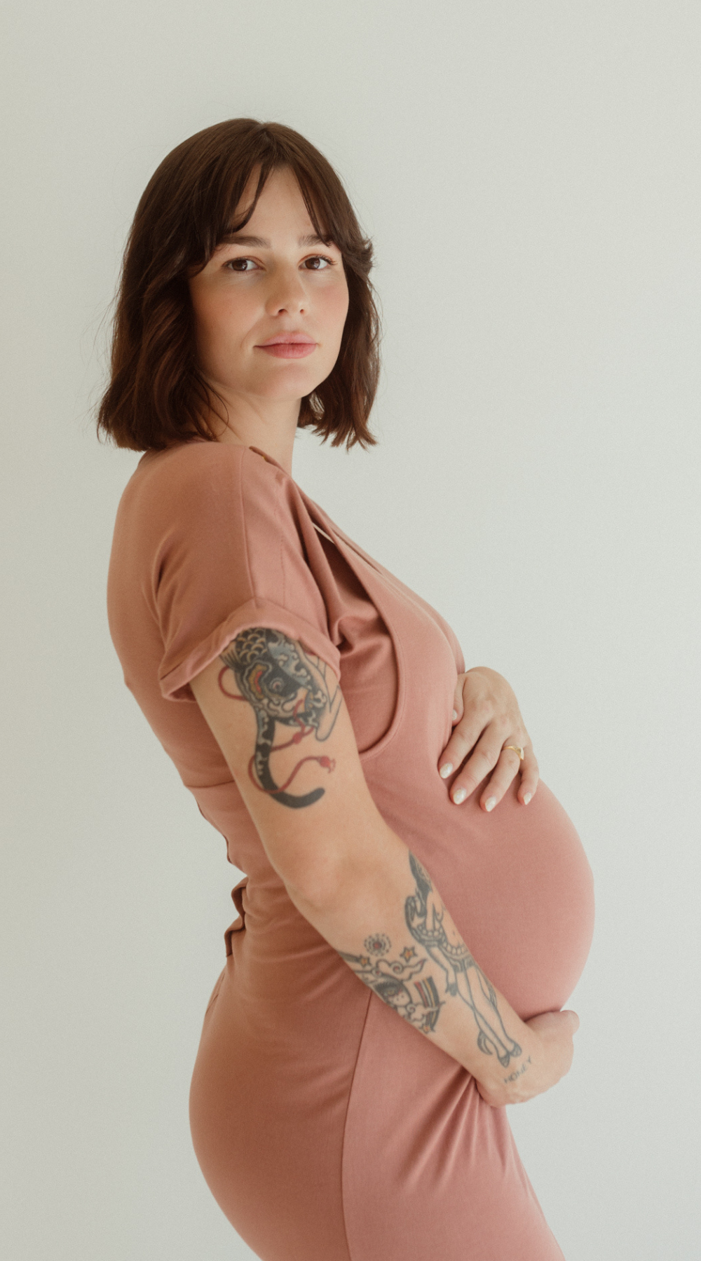 A pregnant mom holds her belly while wearing the Chekoh Journey Dress in the color Dusk - a dusky rose pink tone.