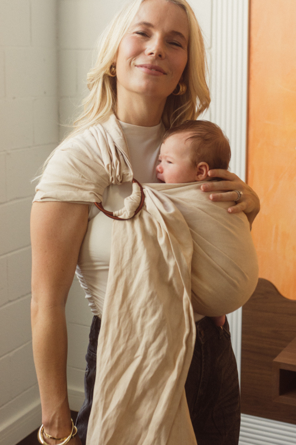 A Brisbane Mum is seen smiling while holding her little newborn close to her chest in the Ring Sling Carrier by Chekoh Baby. It is a soft sandy beige neutral tone with copper rings.