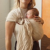 A Brisbane Mum is seen smiling while holding her little newborn close to her chest in the Ring Sling Carrier by Chekoh Baby. It is a soft sandy beige neutral tone with copper rings.