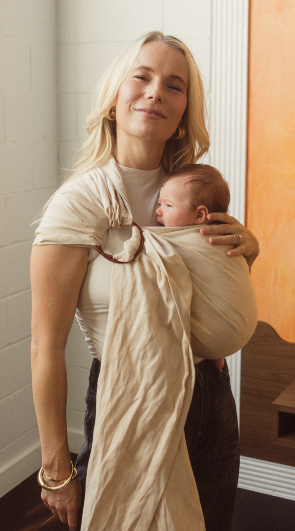 A Brisbane Mum is seen smiling while holding her little newborn close to her chest in the Ring Sling Carrier by Chekoh Baby. It is a soft sandy beige neutral tone with copper rings.