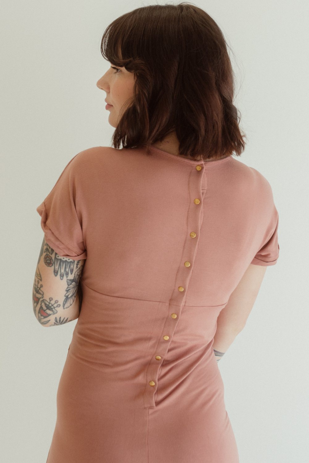 A woman showcases the back details of the Journey Dress by Chekoh in colour Dusk - a dusky pink tone.
