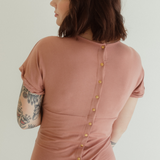 A woman showcases the back details of the Journey Dress by Chekoh in colour Dusk - a dusky pink tone.
