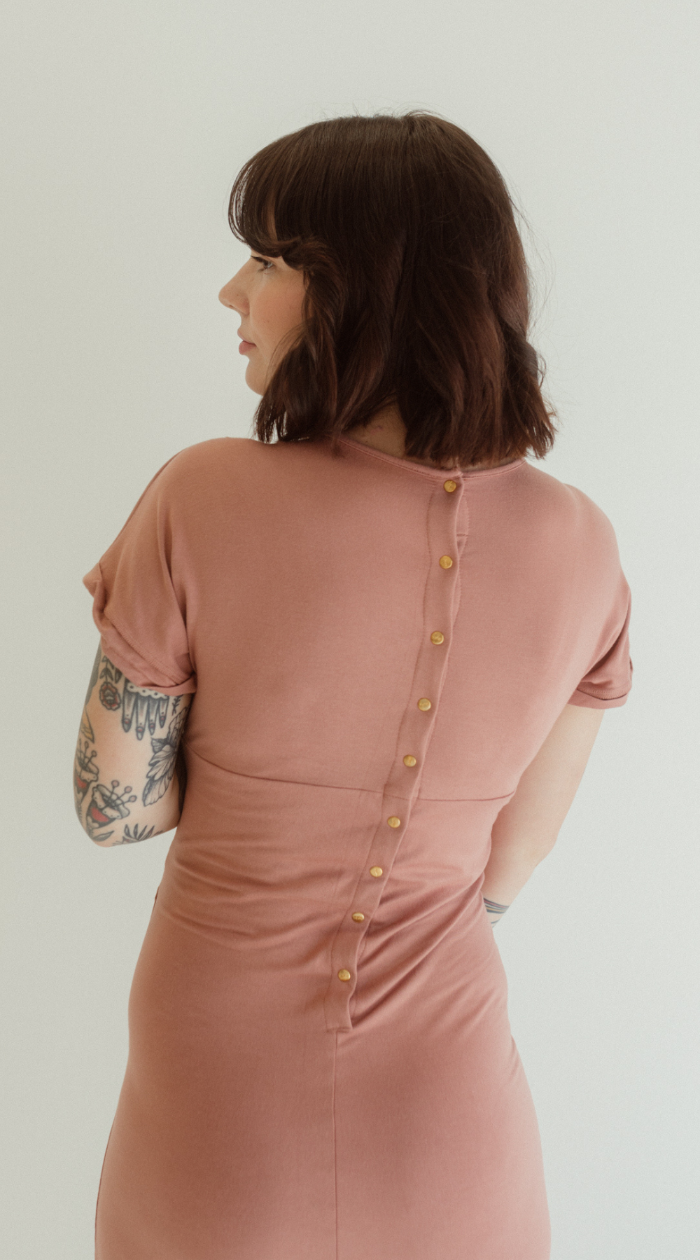 A woman showcases the back details of the Journey Dress by Chekoh in colour Dusk - a dusky pink tone.