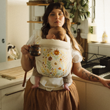 Woman holding a newborn baby in a floral chekoh baby carrier in a kitchen. Juni is a limited edition embroidered folk art Clip Carrier made for all seasons.