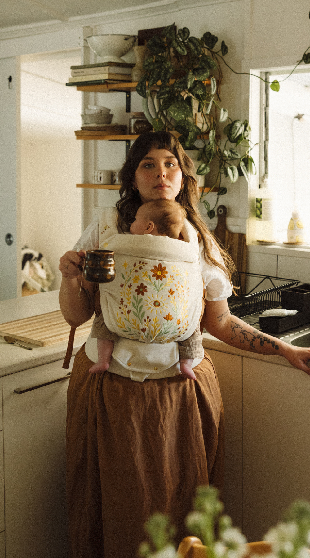 Woman holding a newborn baby in a floral chekoh baby carrier in a kitchen. Juni is a limited edition embroidered folk art Clip Carrier made for all seasons.