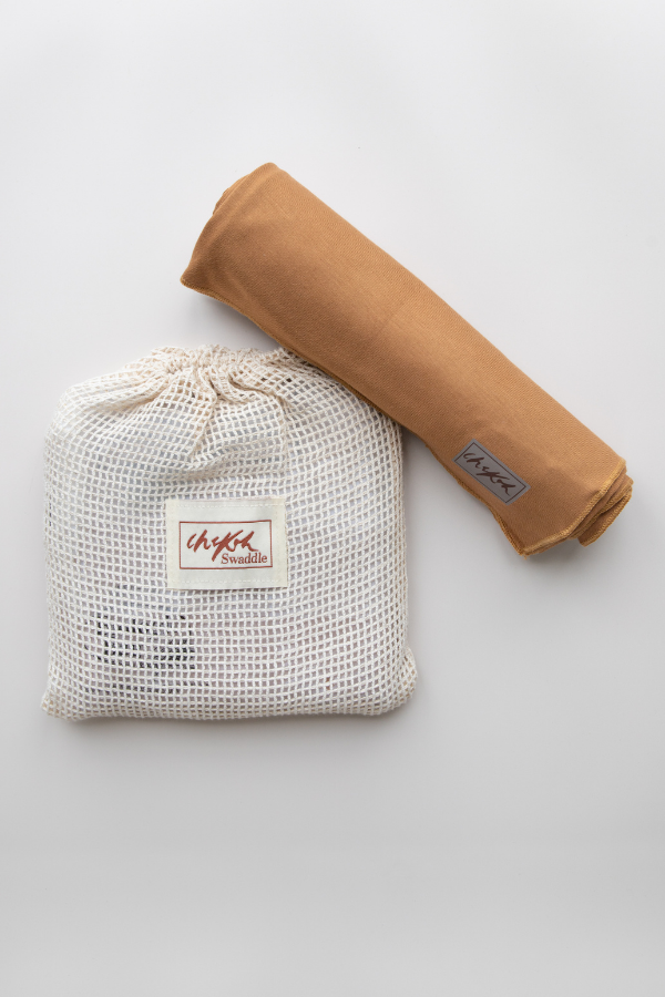 An earthy clay brown Baby Swaddle by Chekoh Baby (Ochre) is sat on a reusable cotton mesh branded wash pouch packaging. 