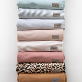 A lineup of all the Chekoh Baby Swaddle Wraps available, Forest (neutral green), Teal, Grey Stripe, Asha (grey), Cinta (blush beige), Cinta Stripe (blush beige stripe on white background), Dusk (now sold out), Leopard (a leopard print exclusive to Chekoh), Ochre (warm earthy brown) and Camel (an earthy golden yellow tone).