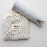 A flatlay image on a white backdrop of the Chekoh Baby Asha Grey Swaddle Wrap. It's soft bamboo stretchy fabric has been rolled for storage and the Swaddle bag is a cotton mesh wash pouch.