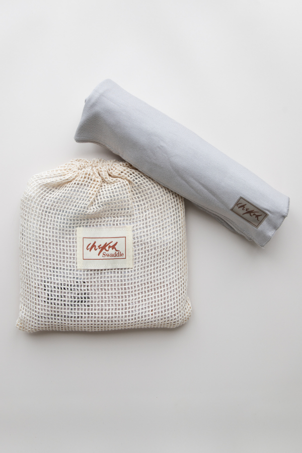 A flatlay image on a white backdrop of the Chekoh Baby Asha Grey Swaddle Wrap. It's soft bamboo stretchy fabric has been rolled for storage and the Swaddle bag is a cotton mesh wash pouch.