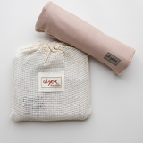 A flatlay image of the Chekoh Baby Cinta Swaddle Stretchy Wrap. It is rolled up and placed on top of the cotton mesh storage pouch/wash bag that it arrives in. Cinta is a blush beige, neutral nude colour.