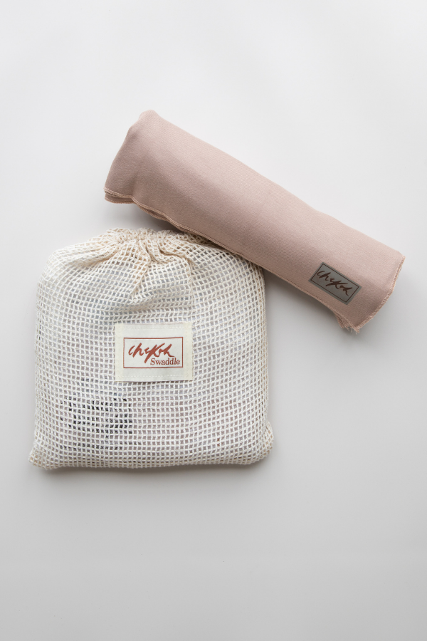 A flatlay image of the Chekoh Baby Cinta Swaddle Stretchy Wrap. It is rolled up and placed on top of the cotton mesh storage pouch/wash bag that it arrives in. Cinta is a blush beige, neutral nude colour.
