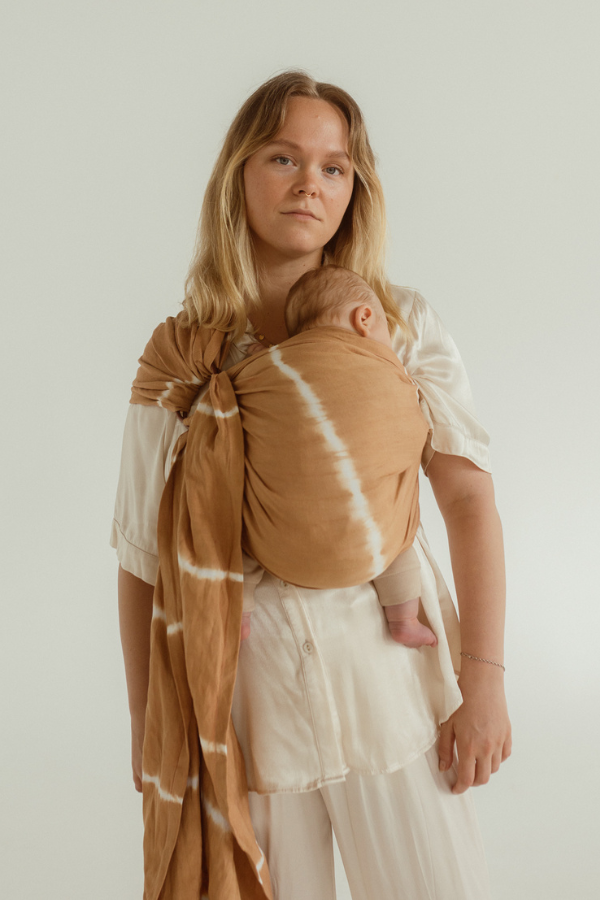A mother is wearing her newborn baby close on her chest with the help of a Chekoh Baby Ring Sling in a caramel brown and white tie dye print. 
