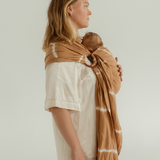 A mother is standing side on, showing the side of the Cove Baby Ring Sling by Chekoh. It is a soft caramel brown tone with white tie dye accents throughout. A perfect Summer neutral paired with an all white outfit.