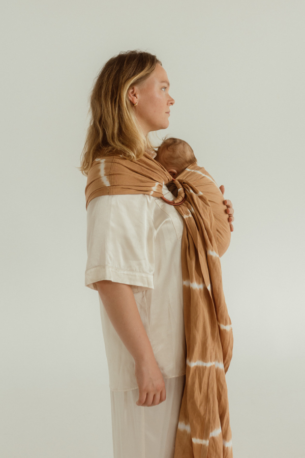 A mother is standing side on, showing the side of the Cove Baby Ring Sling by Chekoh. It is a soft caramel brown tone with white tie dye accents throughout. A perfect Summer neutral paired with an all white outfit.