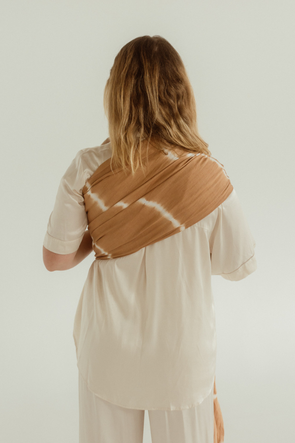 A mother is facing away, showing off the fanned out bamboo/linen back of the baby Ring Sling by Chekoh in the Cove colourway. A caramel brown and white tie dyed print. 