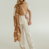 A mother is standing on a white backdrop with her baby in a Ring Sling Carrier by Chekoh Baby. She is wearing an all white satin outfit and the Cove Sling (caramel brown tones with a white contrasting tie dye print) accents beautifully against the plain backdrop.