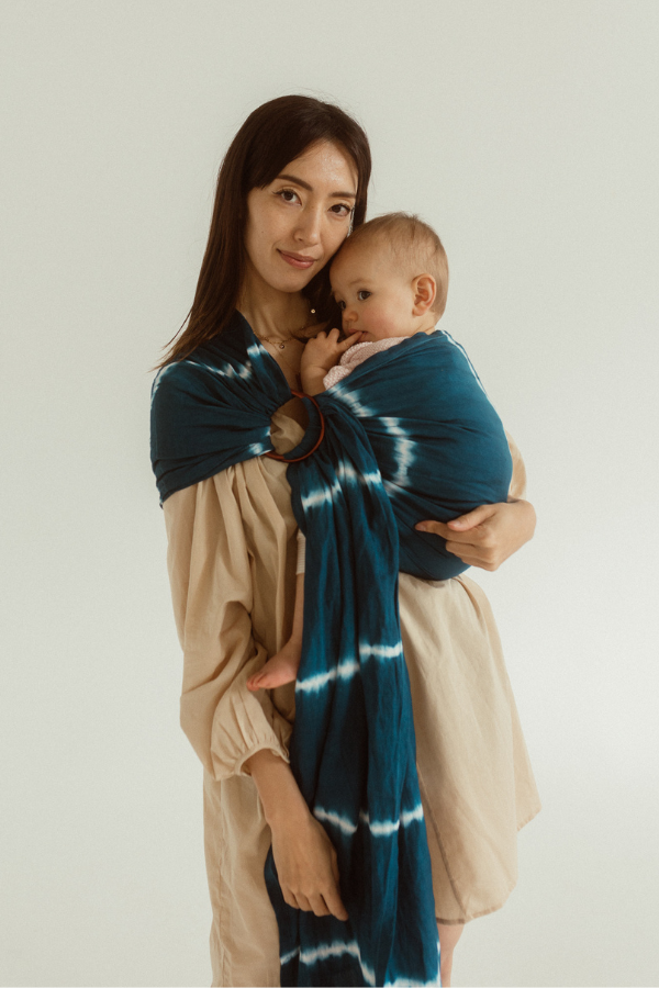 A smiling mother, holds her cute baby close in the Indigo Ring Sling by Chekoh Baby. It features white tie-dyed lines on a deep indigo colours background. 