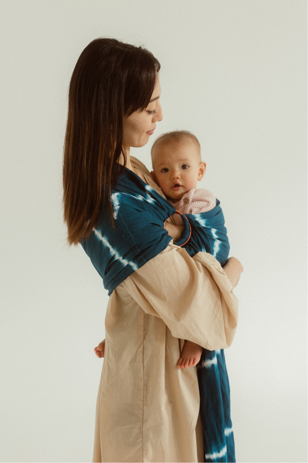 A cute baby with fluffy hair is cuddled close in a Chekoh Baby Ring Sling in the tie-dyed print 'Indigo'. 