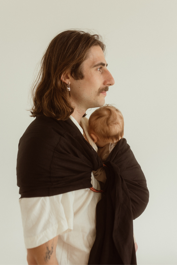 A dad and baby are facing side on, revealing small details of the Chekoh Baby Ring Sling Carrier, including the copper coloured rings and linen/bamboo blend fabric.