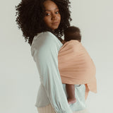 A mother and newborn stand side on while wearing the Chekoh baby Ring Sling in Terracotta, a light beige/pink neutral toned option for parents.