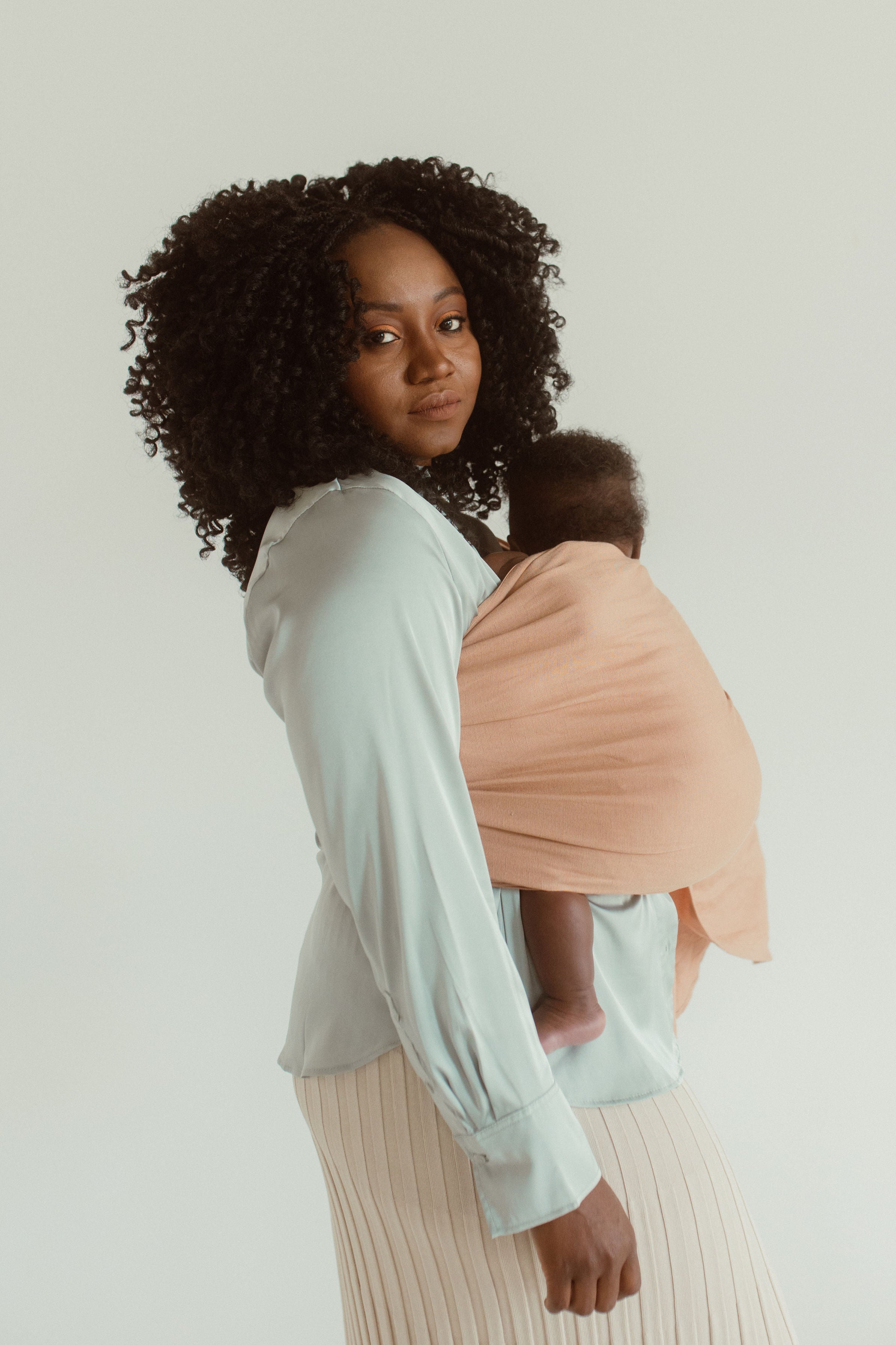 A mother and newborn stand side on while wearing the Chekoh baby Ring Sling in Terracotta, a light beige/pink neutral toned option for parents.