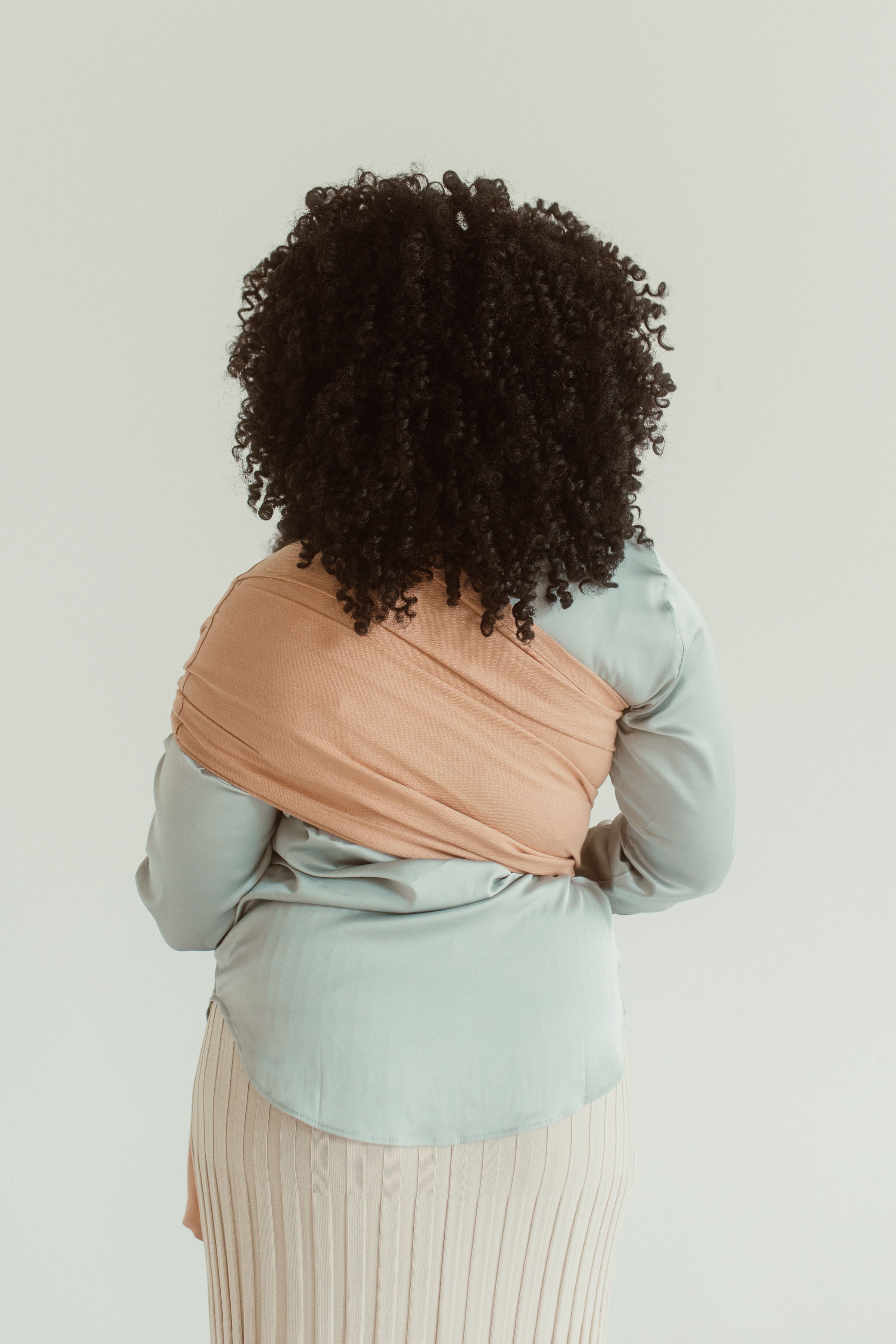 A mother faces away to show off the back view of a Chekoh baby Ring Sling Carrier. It is seen over her shoulder, made from linen and bamboo in a Terracotta shade. 