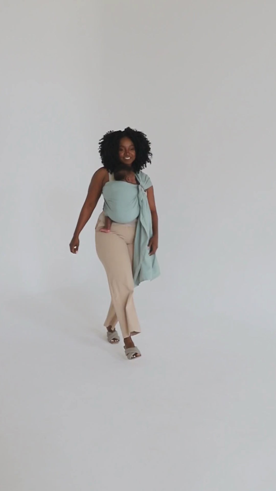A video of a mother, wearing her baby in a ring sling carrier by Chekoh baby, walking across a white background. 