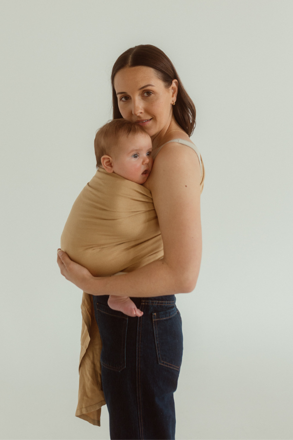 A cute baby is close to their mother inside of a Chekoh Baby Ring Sling Carrier in the colour Camel - a soft yellow tone.
