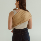 A mother and baby are facing away, showing the back of the Ring Sling Carrier by Chekoh - a soft yellow toned camel colourway fanned evenly across one shoulder and opposite side waist. 